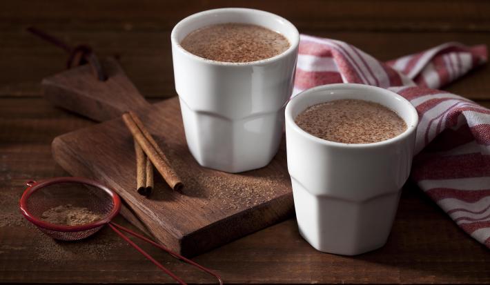 Foto dois copos brancos com chocolate quente dentro em cima de uma tábua de madeira com canela em pau e em pó decorando a mesa